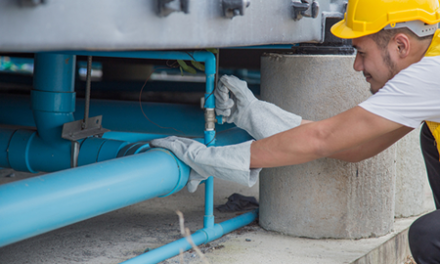 worker in hard hat repairs water pipe line under building